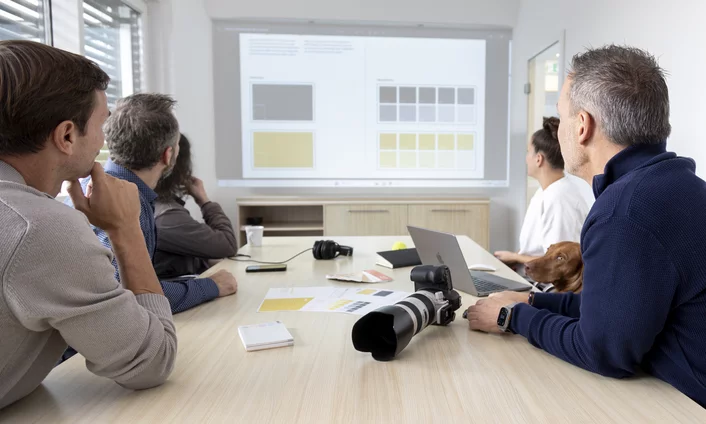 Team im Meeting an einem Besprechungstisch mit Kamera und Laptop, auf der Leinwand Entwürfe für ein Farbkonzept