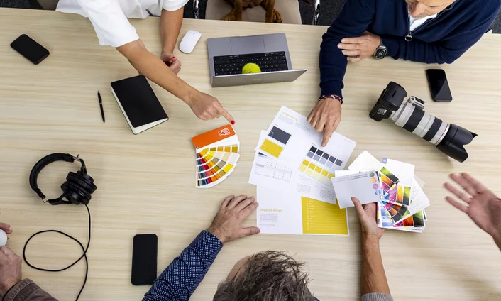 Werte & Haltung Team bespricht am Tisch Farbmuster und Corporate-Design-Entwürfe, daneben Laptop, Kamera und Kopfhörer