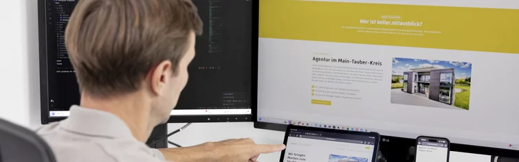 Mitarbeiter zeigt an einem Arbeitsplatz mit mehreren Bildschirmen auf eine Website auf dem Tablet